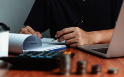 Ein Arbeitsplatz mit Dokumenten, einem Taschenrechner, einem Laptop und Münzen im Vordergrund